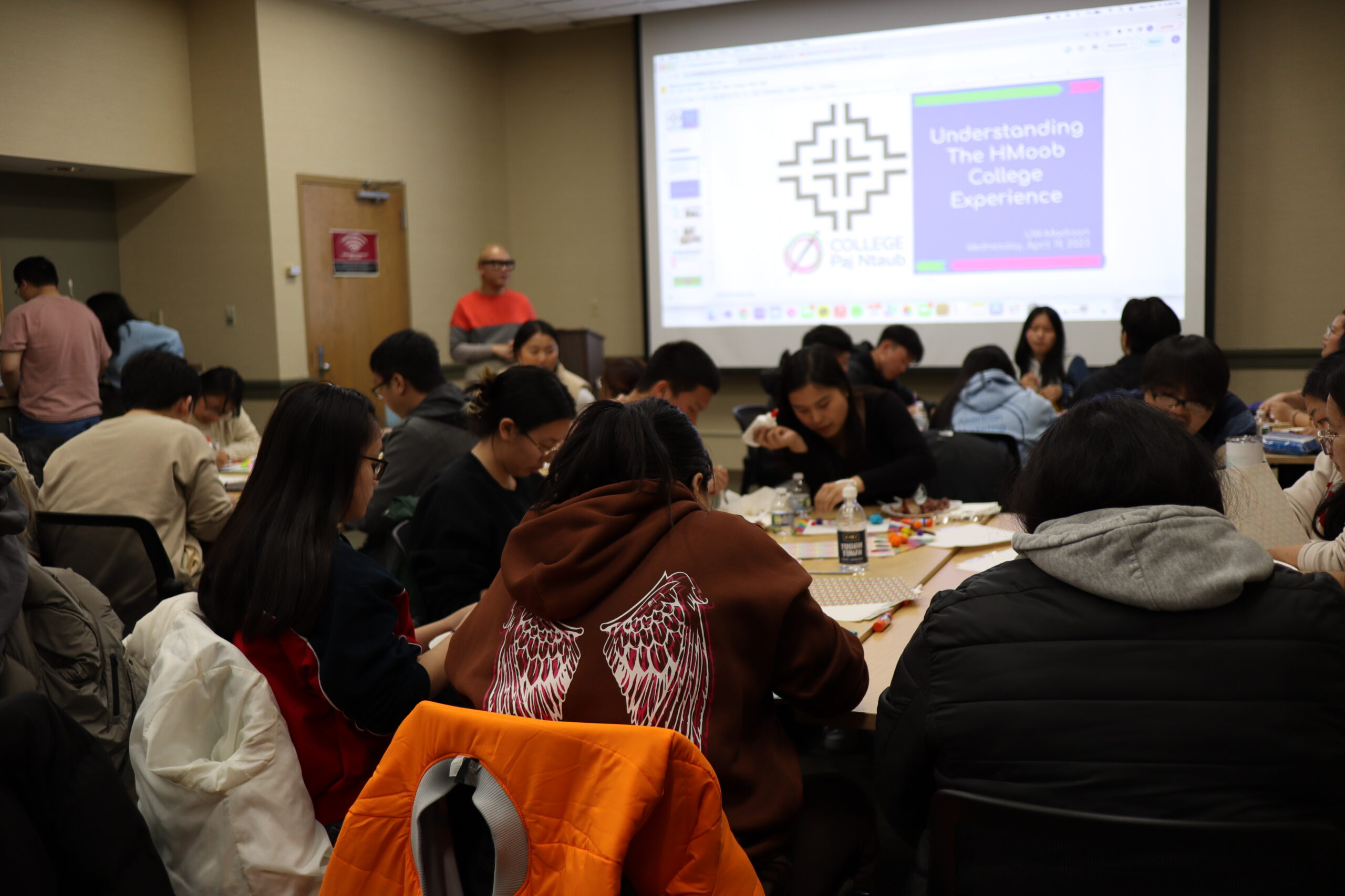 Room of students sitting at tables with a projection screen in the background.