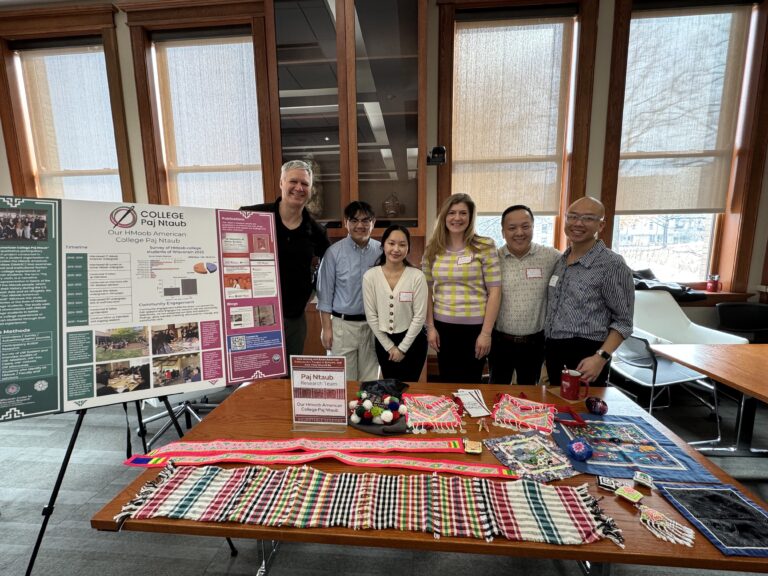 College Paj Ntaub research team standing at a table with items displayed on it with a poster on an easel next to them.