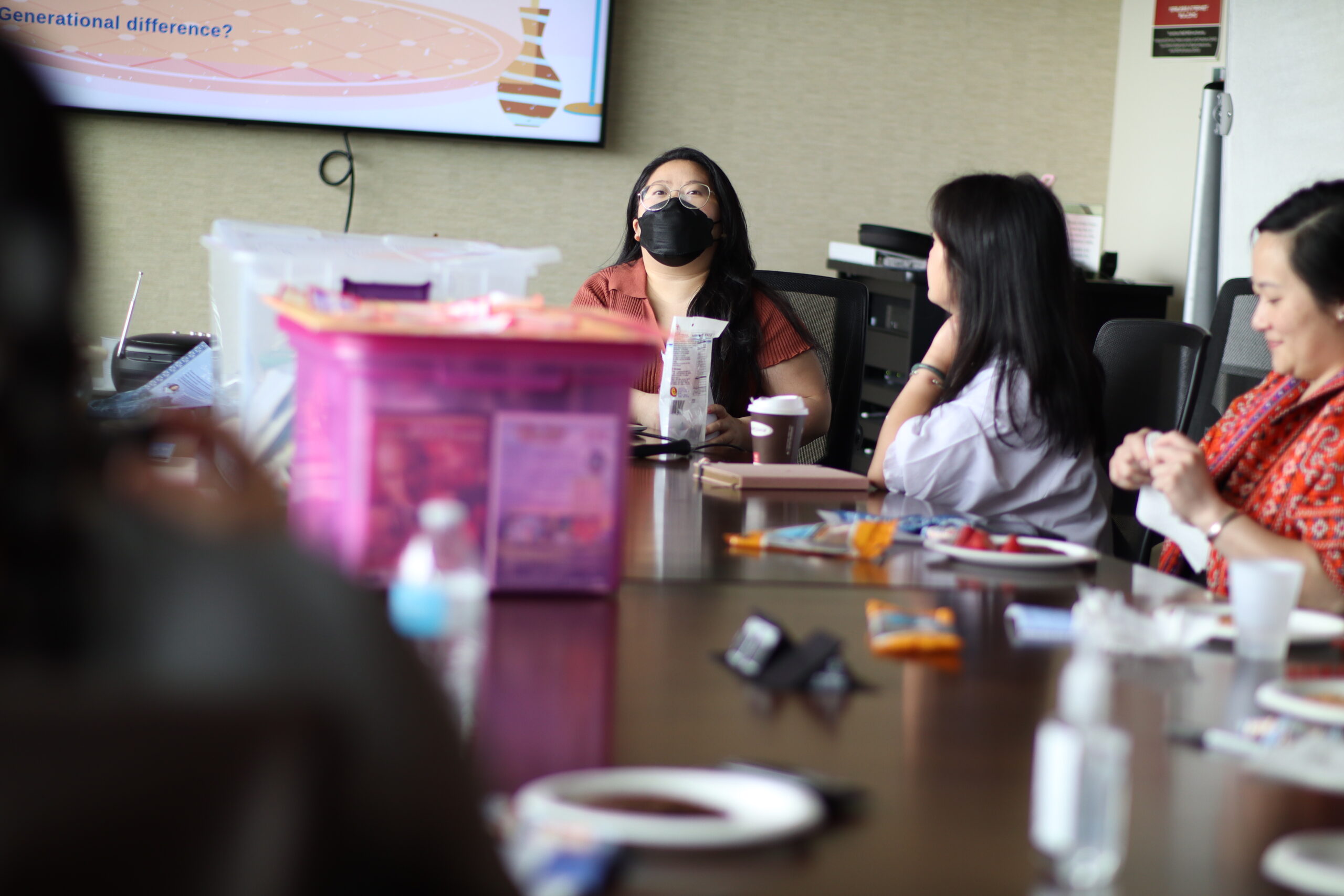 A presenter wearing a mask is looking across a table with various items sitting on it. Two other people sitting at the table.