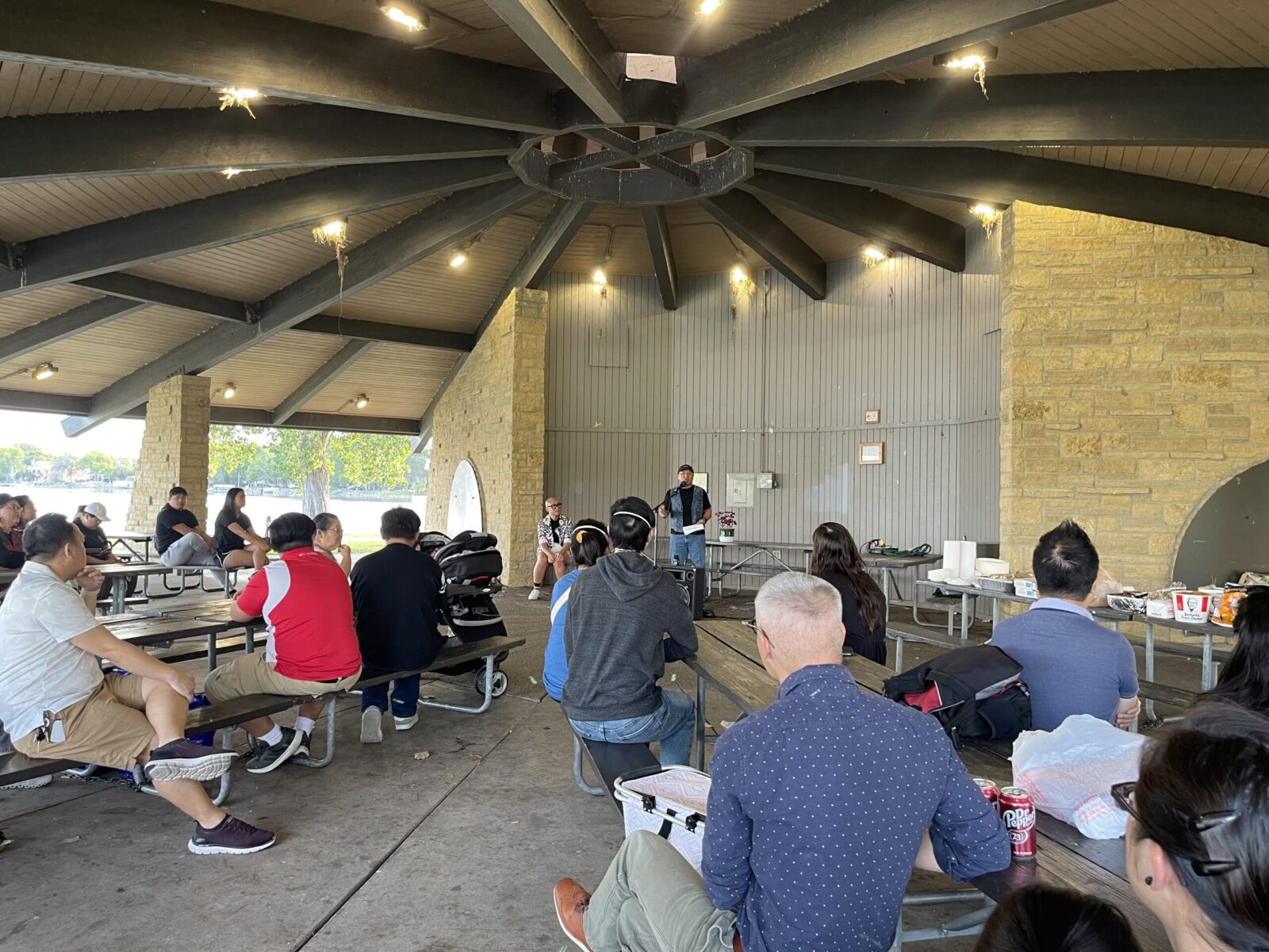 Speaker presenting to a group sitting at picnic tables in an outdoor park shelter.